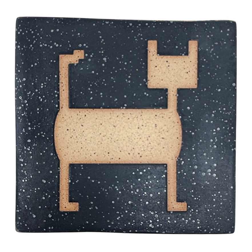 A matte black ceramic tile with a shape of a cat on a speckled stoneware background.
